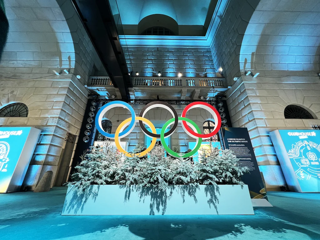 Anelli olimpici sopra una fioriera di abeti innevati in una sala di pietra immersa in una gelida luce blu – Clubhouse 26 Milano, Italia, 2026 – hospitality design