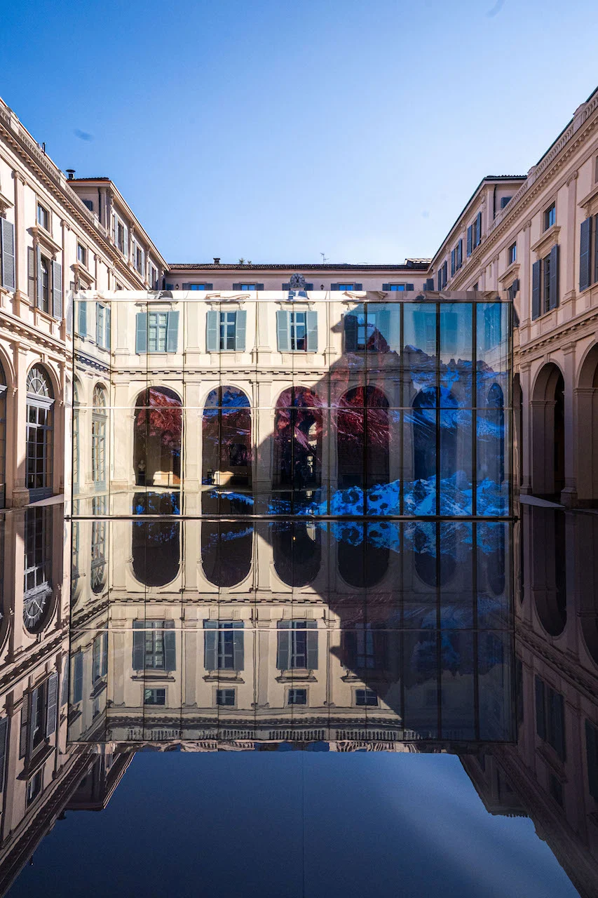 Padiglione di vetro con l'immagine di una montagna innevata, riflesso con i portici del cortile nella vasca scura – Samsung House a Palazzo Serbelloni, Milano, Italia, 2026 – interior design
