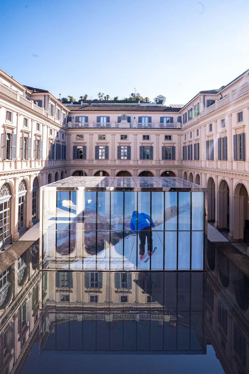 Padiglione di vetro nel cortile con uno sciatore su un maxischermo, riflesso nella vasca nera – Samsung House a Palazzo Serbelloni, Milano, Italia, 2026 – interior design