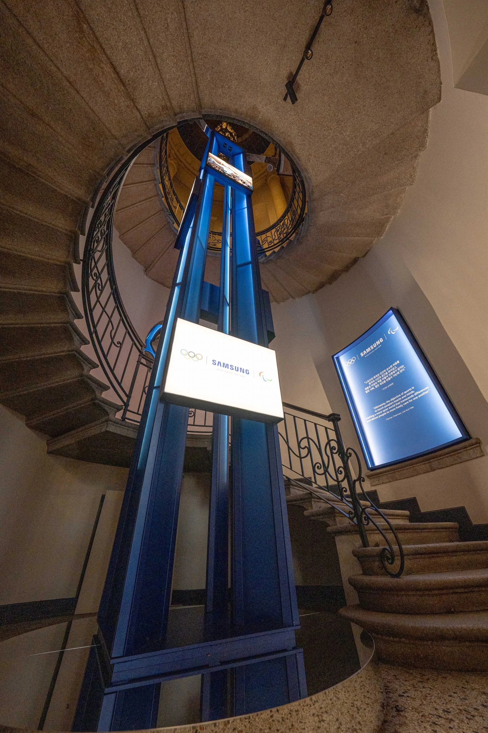 Vano ascensore in acciaio blu con insegne Samsung e olimpiche che sale attraverso una scala elicoidale in pietra – Samsung House a Palazzo Serbelloni, Milano, Italia, 2026 – interior design