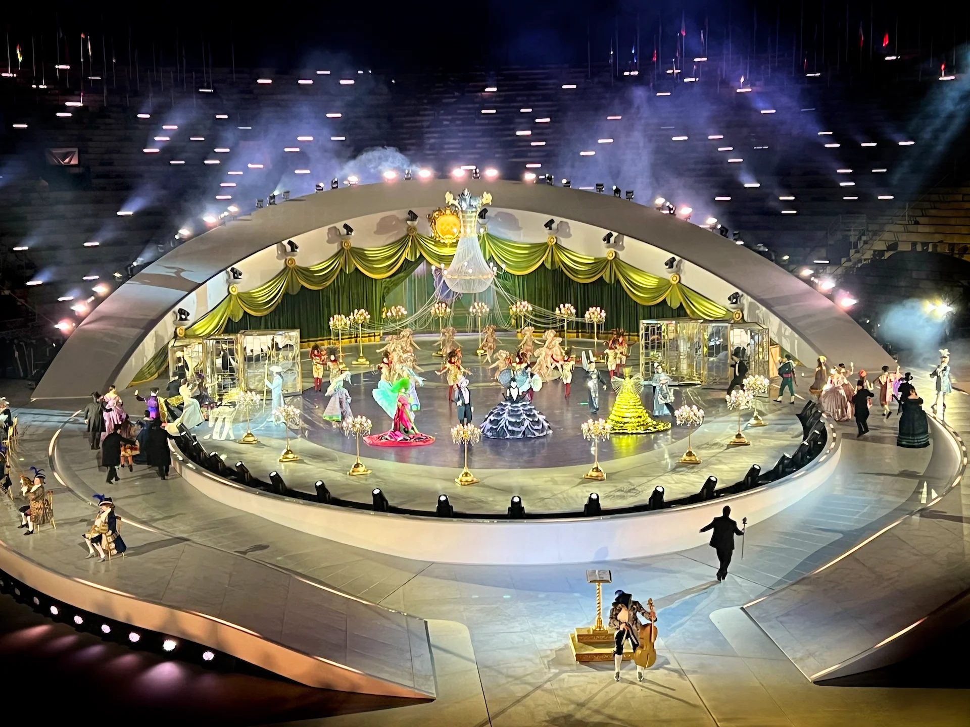 Artisti in costumi e abiti in stile barocco sul palco centrale, incorniciati da drappi verdi e oro – Cerimonia di chiusura dei Giochi Olimpici di Milano Cortina 2026, Verona, Italia – scenografia