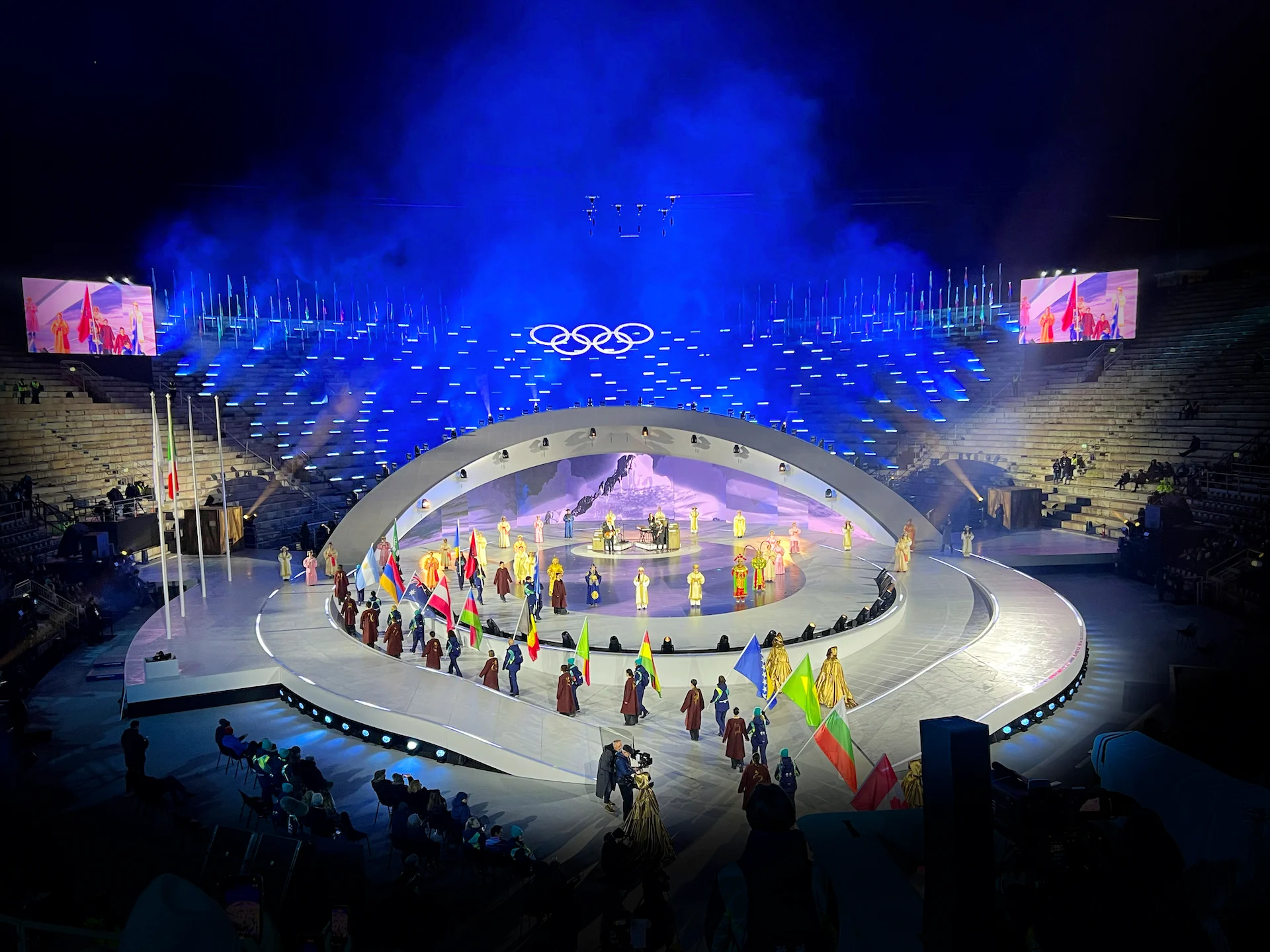 Portabandiera in parata attorno al palco ovale sotto gli anelli olimpici, foschia blu sulle gradinate dell'arena – Cerimonia di chiusura dei Giochi Olimpici di Milano Cortina 2026, Verona, Italia – scenografia