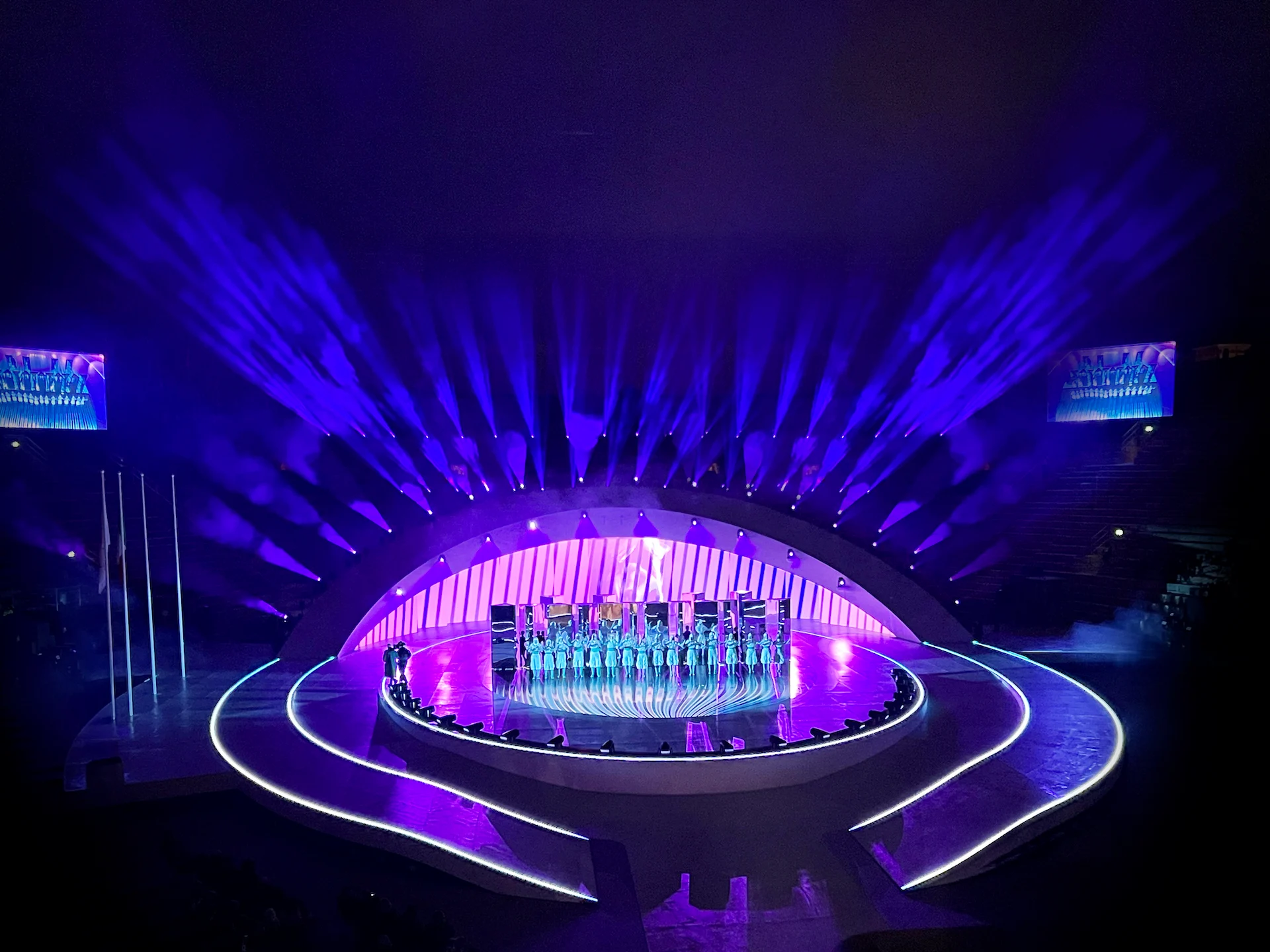 Coro sul palco centrale sotto l'arco curvo, incorniciato da un ventaglio di fasci di luce viola – Cerimonia di chiusura dei Giochi Olimpici di Milano Cortina 2026, Verona, Italia – scenografia