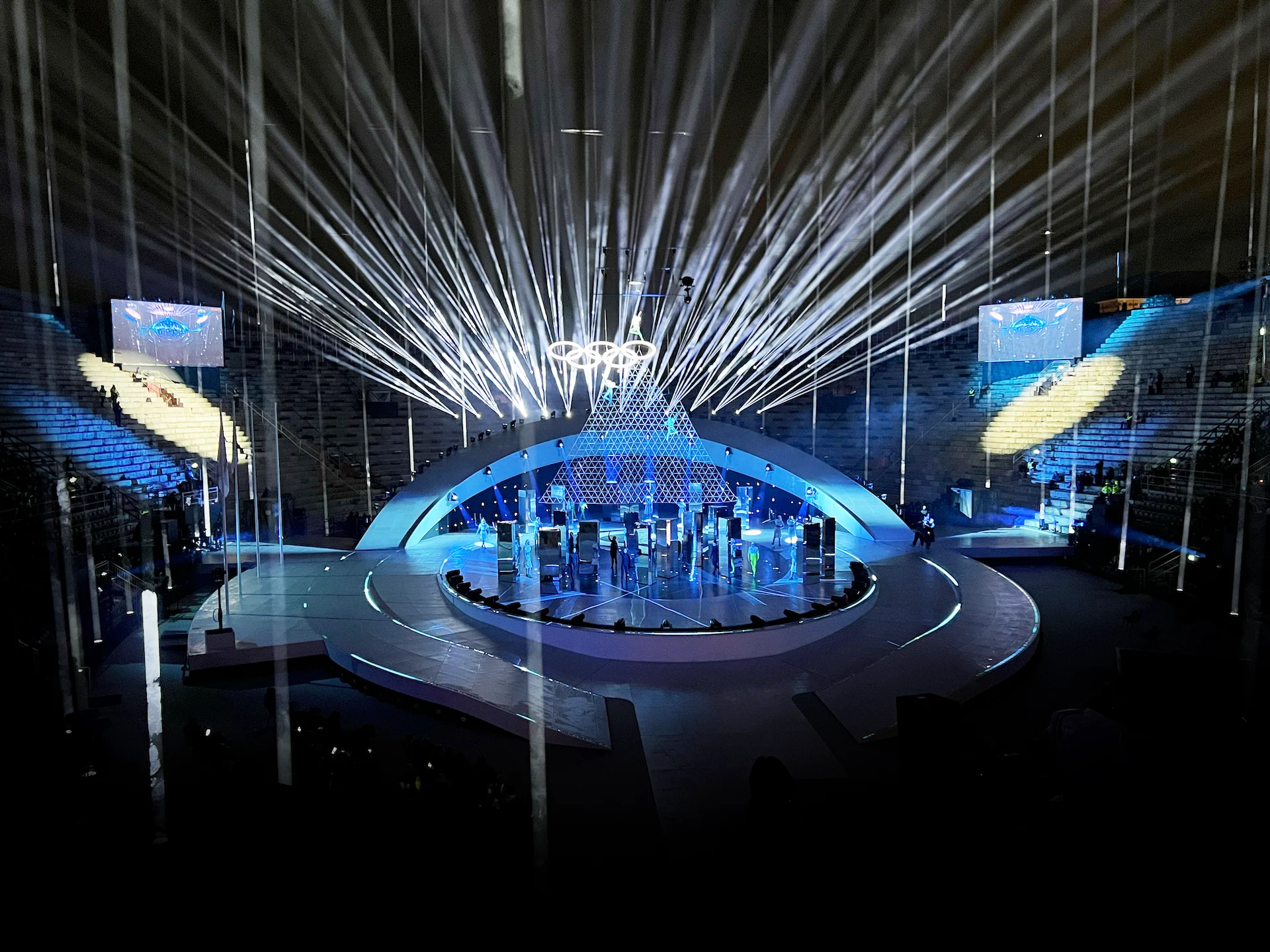 Vista d'insieme del palco della cerimonia di chiusura all'Arena di Verona: pedana ovale sotto un arco curvo e fasci di luce bianca – Cerimonia di chiusura dei Giochi Olimpici di Milano Cortina 2026, Verona, Italia – scenografia