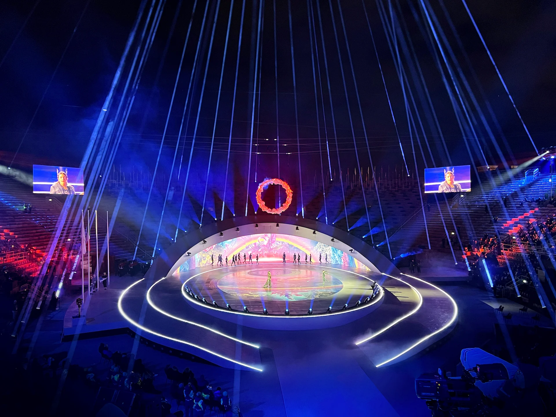 Artisti sul palco illuminato sotto un anello di fiamme, con fasci di luce che scendono dall'alto – Cerimonia di chiusura dei Giochi Olimpici di Milano Cortina 2026, Verona, Italia – scenografia