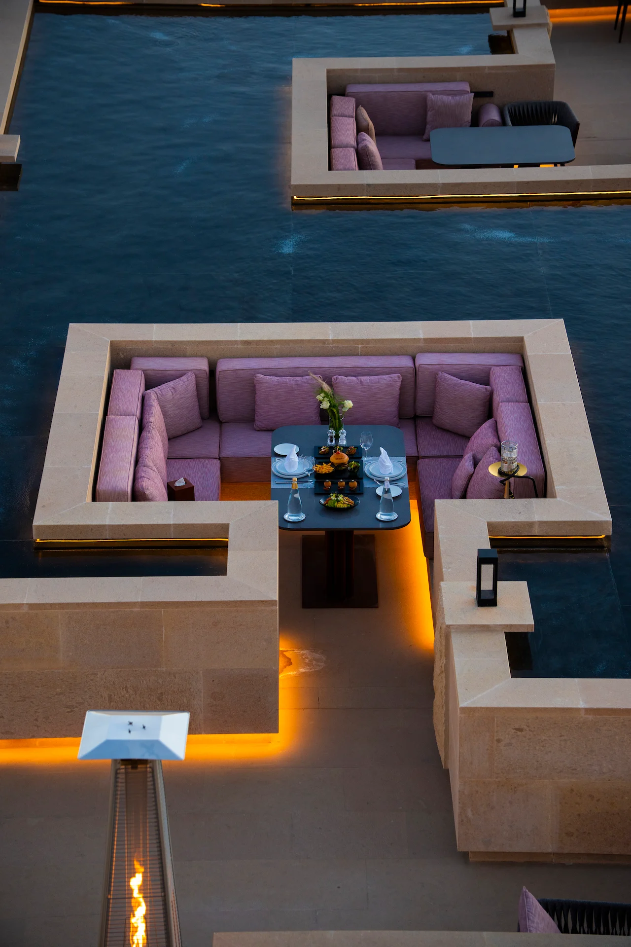 Salottino ribassato con cuscini rosa e una tavola apparecchiata, circondato dall'acqua nella vasca di pietra – Ristorante Sky View presso The Chedi Hegra, AlUla, Arabia Saudita, 2026 – architettura