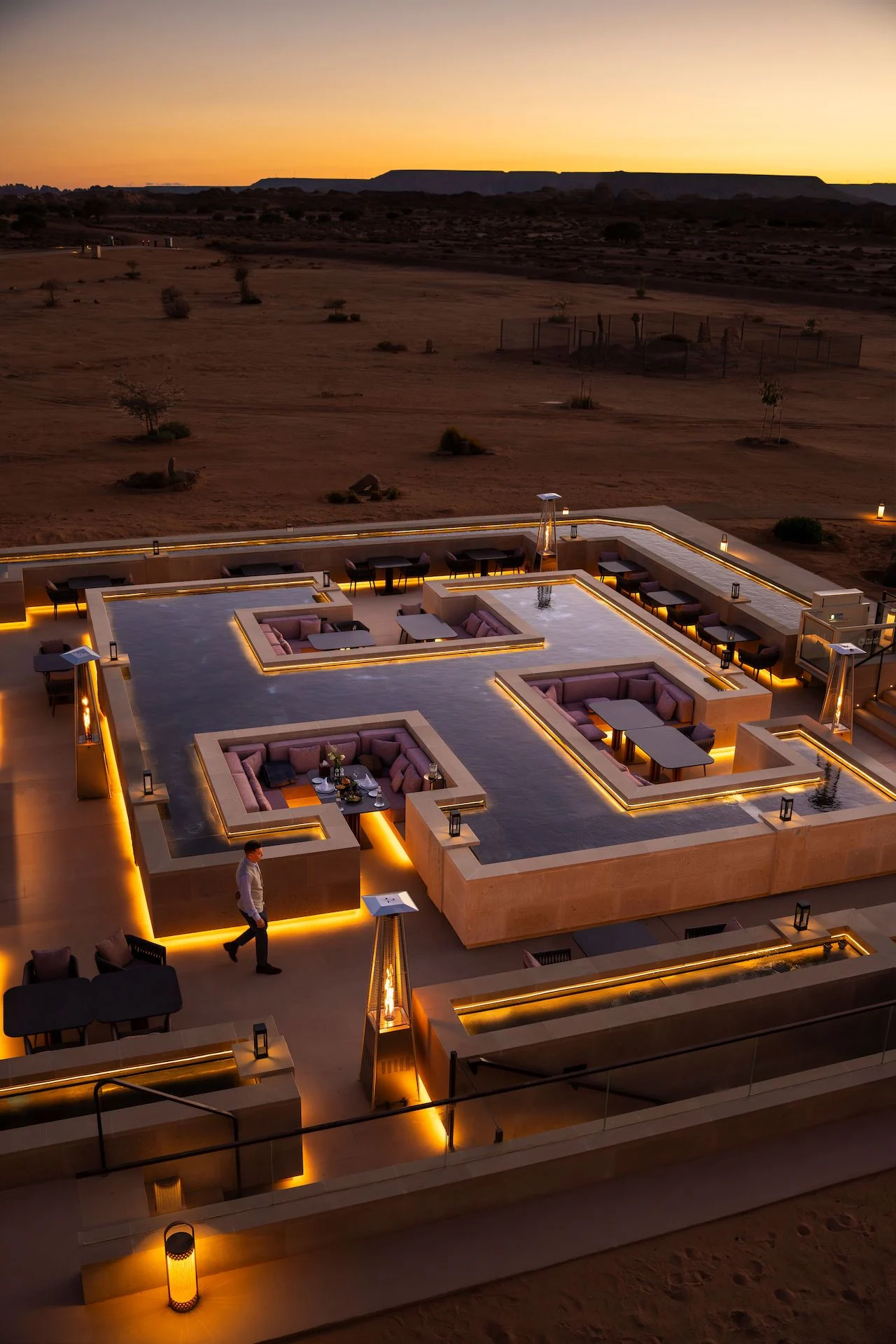 Sky View Restaurant al crepuscolo dall'alto: salotti ribassati in una vasca scura a specchio, il deserto sullo sfondo – Ristorante Sky View presso The Chedi Hegra, AlUla, Arabia Saudita, 2026 – architettura