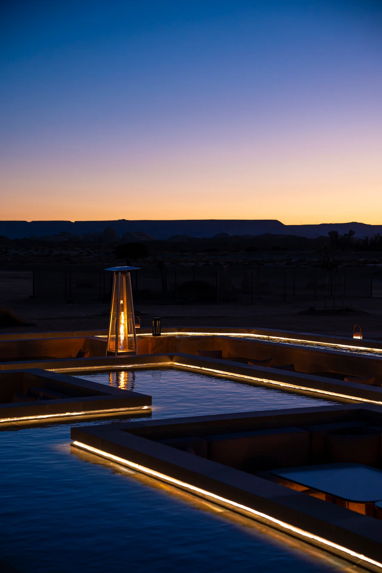 Fungo riscaldante acceso e bordi della vasca illuminati al crepuscolo, con altopiani desertici all'orizzonte – Ristorante Sky View presso The Chedi Hegra, AlUla, Arabia Saudita, 2026 – architettura