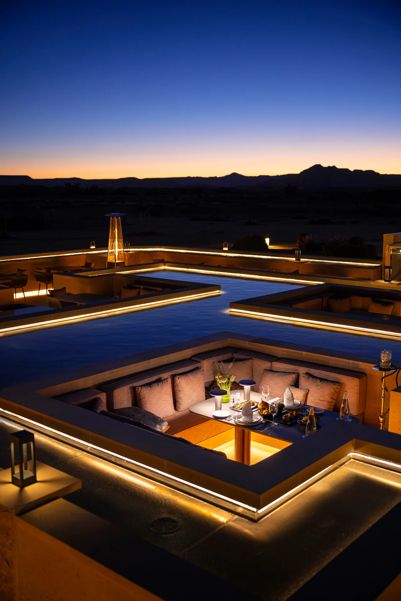 Salottino ribassato con un tavolo apparecchiato per la cena all'ora blu, linee di luce lungo i bordi della vasca – Ristorante Sky View presso The Chedi Hegra, AlUla, Arabia Saudita, 2026 – architettura