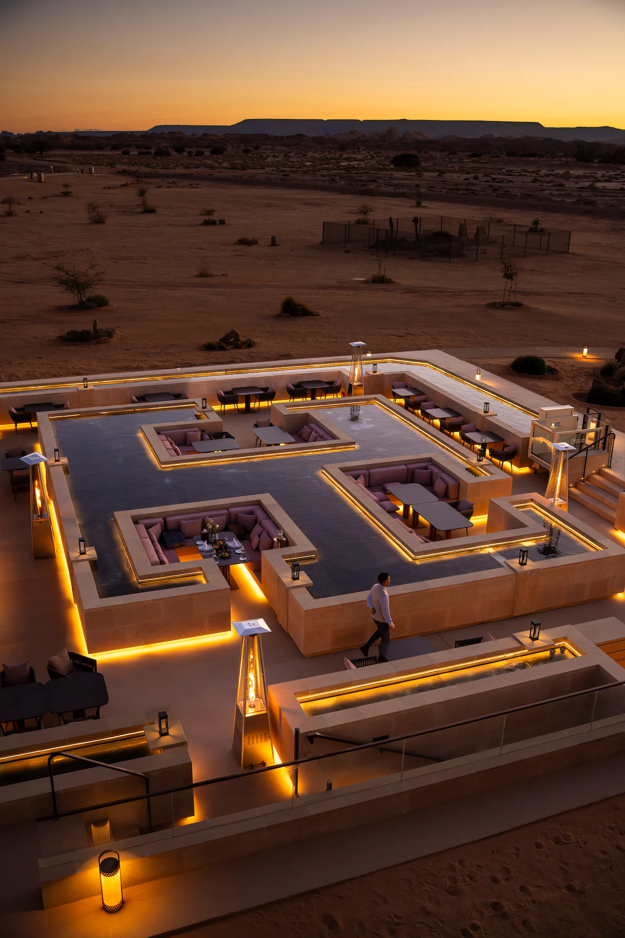 Vista dall'alto della terrazza del ristorante al tramonto, un cameriere che passa tra i funghi riscaldanti, la pianura desertica dietro – Ristorante Sky View presso The Chedi Hegra, AlUla, Arabia Saudita, 2026 – architettura
