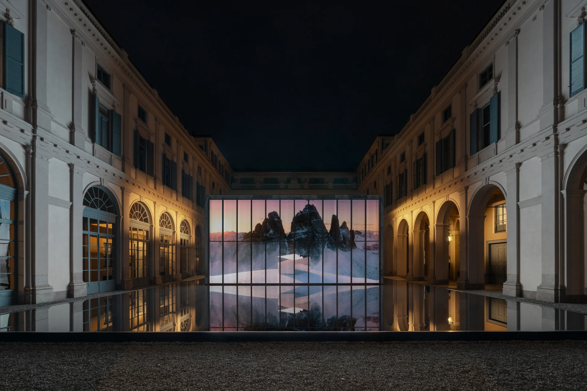 Cortile di notte: padiglione di vetro illuminato con l'immagine di aspre vette montuose, riflesso nella vasca nera – Samsung House a Palazzo Serbelloni, Milano, Italia, 2026 – interior design