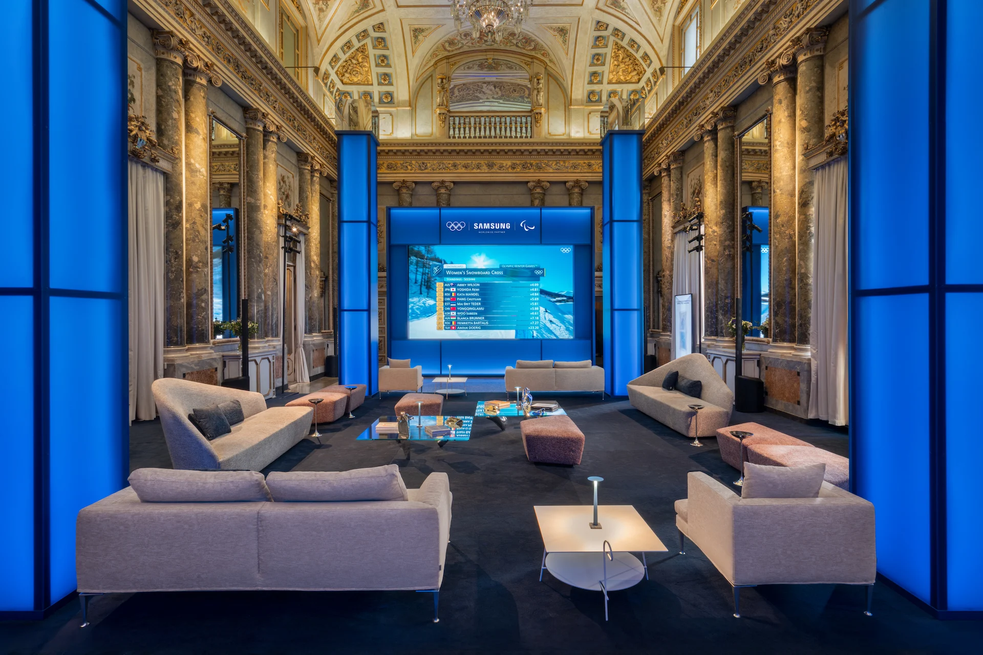 Salone affrescato del palazzo con colonne di luce blu, divani chiari e un grande schermo con i risultati olimpici – Samsung House a Palazzo Serbelloni, Milano, Italia, 2026 – interior design