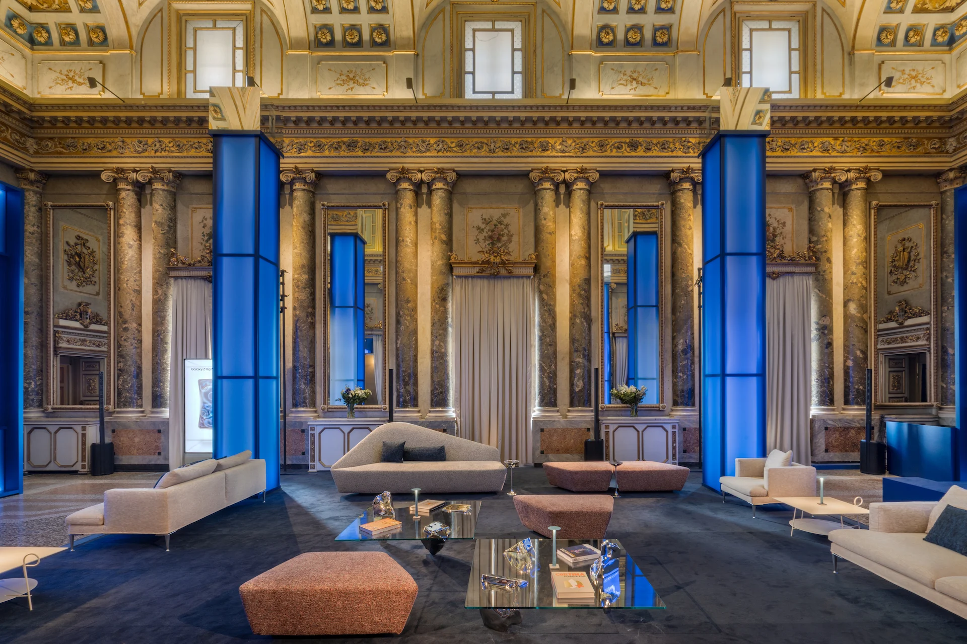 Salone dorato con colonne di marmo e pilastri illuminati di blu, arredato con divani chiari, tavolini di vetro e pouf rosa – Samsung House a Palazzo Serbelloni, Milano, Italia, 2026 – interior design