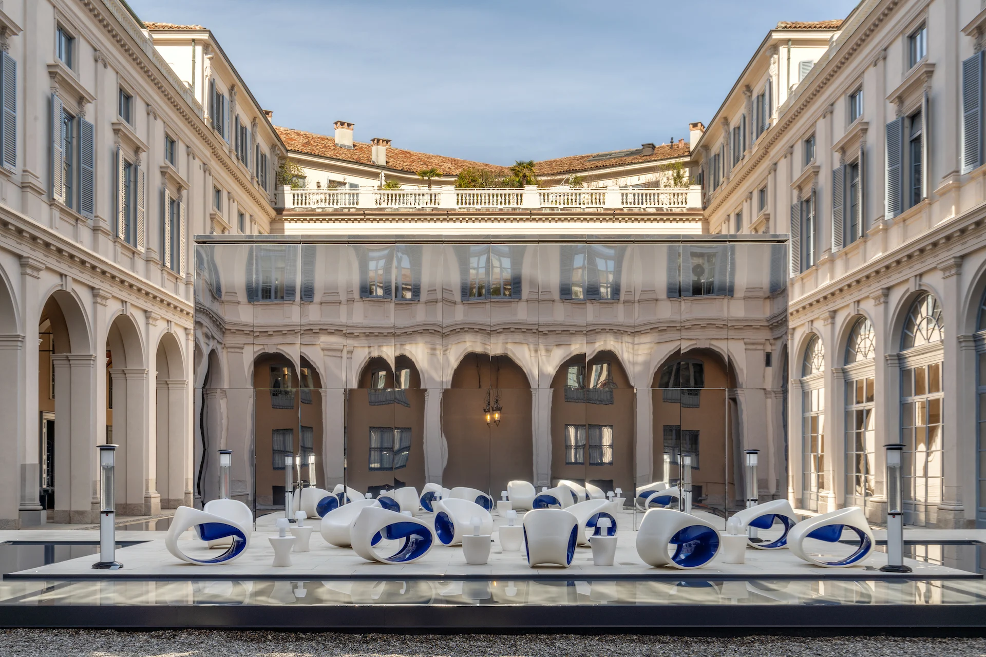 Sedute lounge bianche e blu su una pedana riflettente nel cortile, sullo sfondo il padiglione di vetro e i portici – Samsung House a Palazzo Serbelloni, Milano, Italia, 2026 – interior design