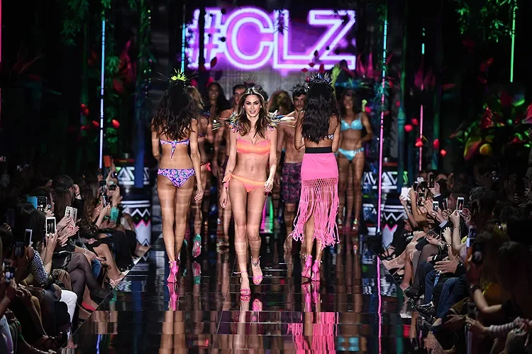 Modelle in costume da bagno sulla passerella a specchio verso il pubblico, sotto l'insegna al neon rosa #CLZ – Collezione Calzedonia SS15, Italia, 2015 – scenografia per sfilate