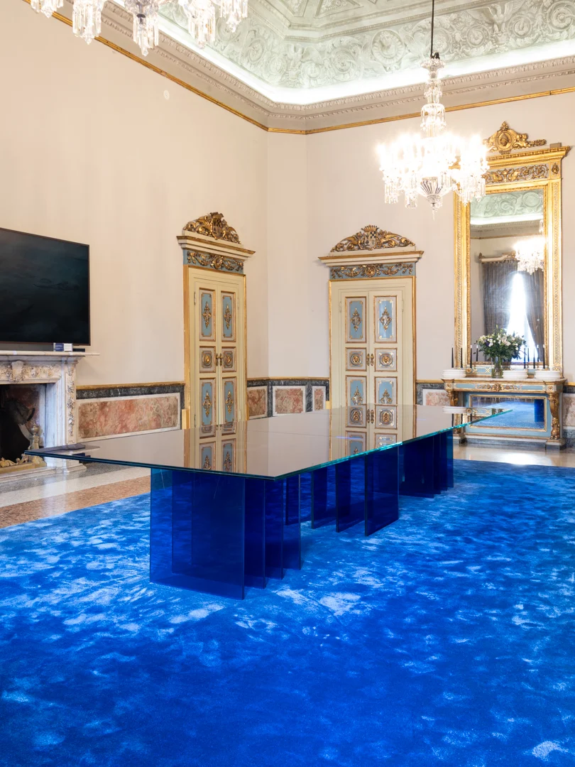 Tavolo STRACTA con gambe in vetro blu stratificato su un tappeto blu, in una sala del palazzo con porte dorate – Tavolo STRACTA per Driade, Milano, Italia, 2026 – design di prodotto