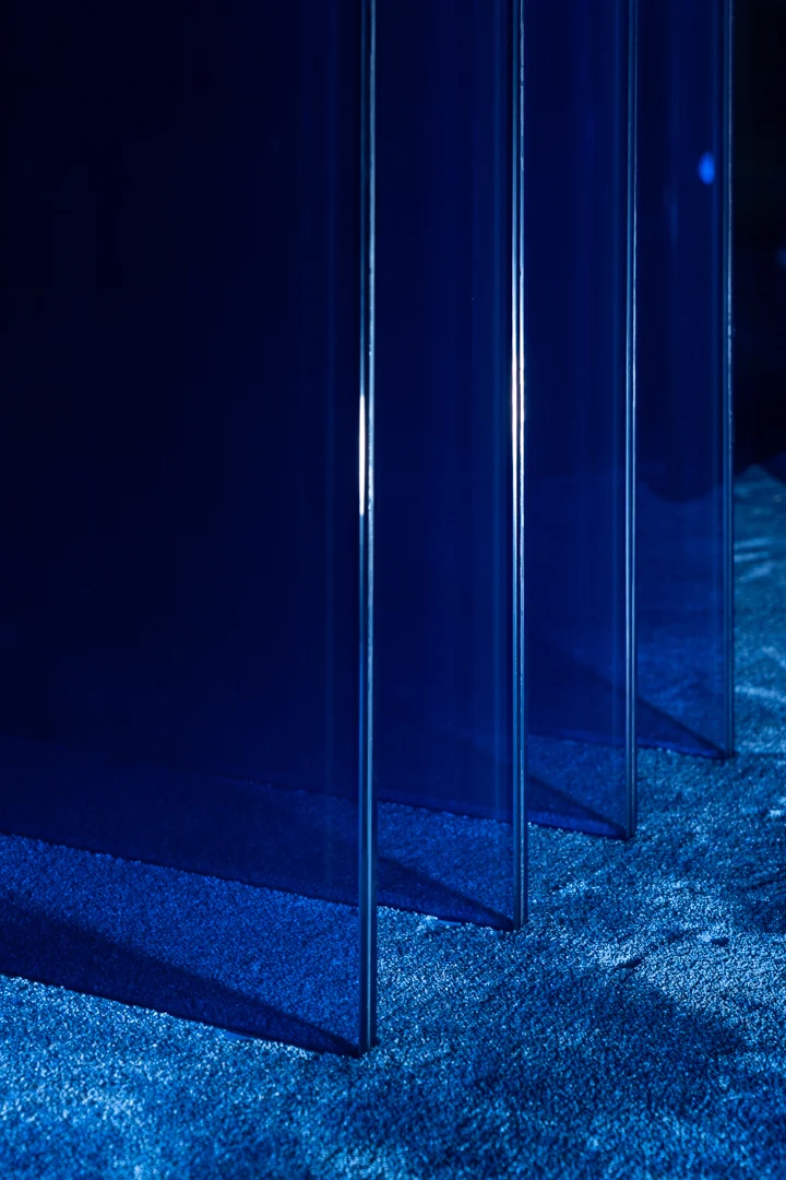 Dettaglio dei pannelli in vetro blu della base che proiettano ombre sulla moquette blu – Tavolo STRACTA per Driade, Milano, Italia, 2026 – design di prodotto