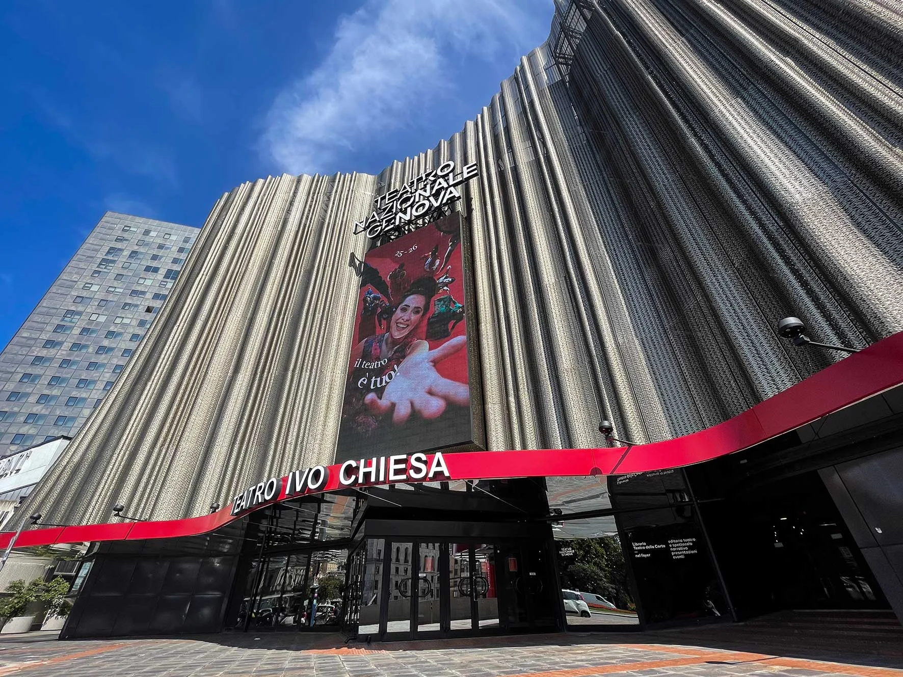 Vista dal basso della facciata del Teatro Nazionale Ivo Chiesa: una cortina metallica plissettata sopra una pensilina d'ingresso rossa – Facciata del Teatro Nazionale Ivo Chiesa, Genova, Italia, 2025 – riqualificazione architettonica