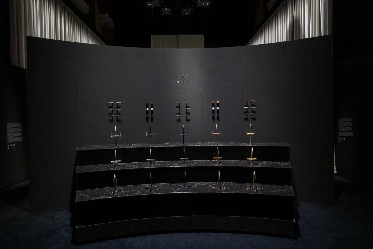 Parete espositiva curva e scura con file di rubinetti in diverse finiture su piani a gradoni in pietra nera – GROHE SPA Aqua Sanctuary al Fuorisalone, Milano, Italia, 2026 – progettazione di installazioni