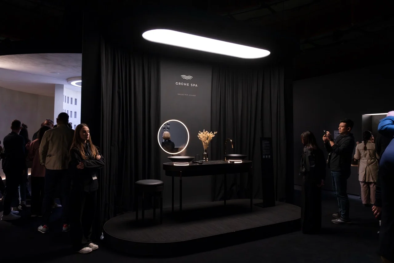 Mobile lavabo scuro con uno specchio rotondo illuminato sotto una luce ovale, visitatori intorno al box con tende – GROHE SPA Aqua Sanctuary al Fuorisalone, Milano, Italia, 2026 – progettazione di installazioni