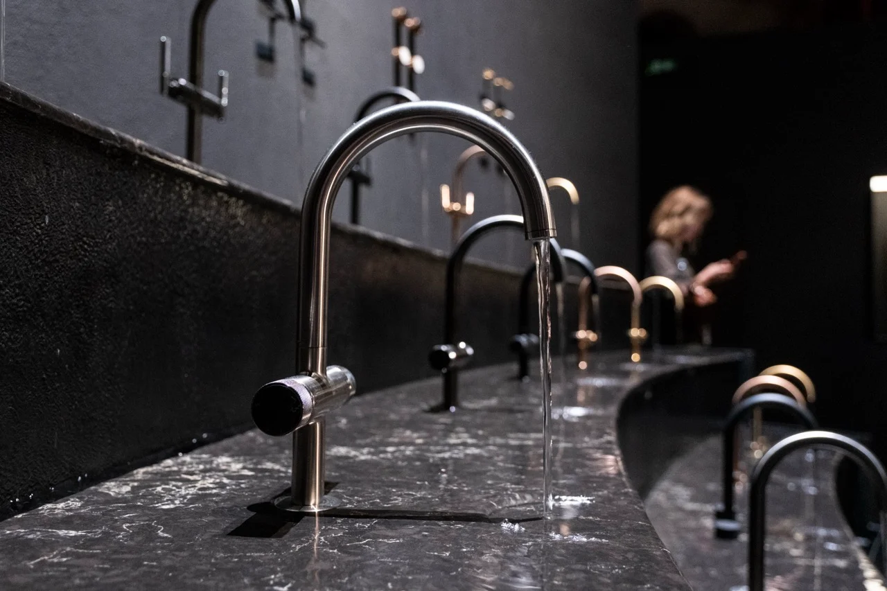 Dettaglio di un rubinetto d'acciaio con acqua corrente su un piano curvo in marmo scuro con una fila di rubinetti – GROHE SPA Aqua Sanctuary al Fuorisalone, Milano, Italia, 2026 – progettazione di installazioni