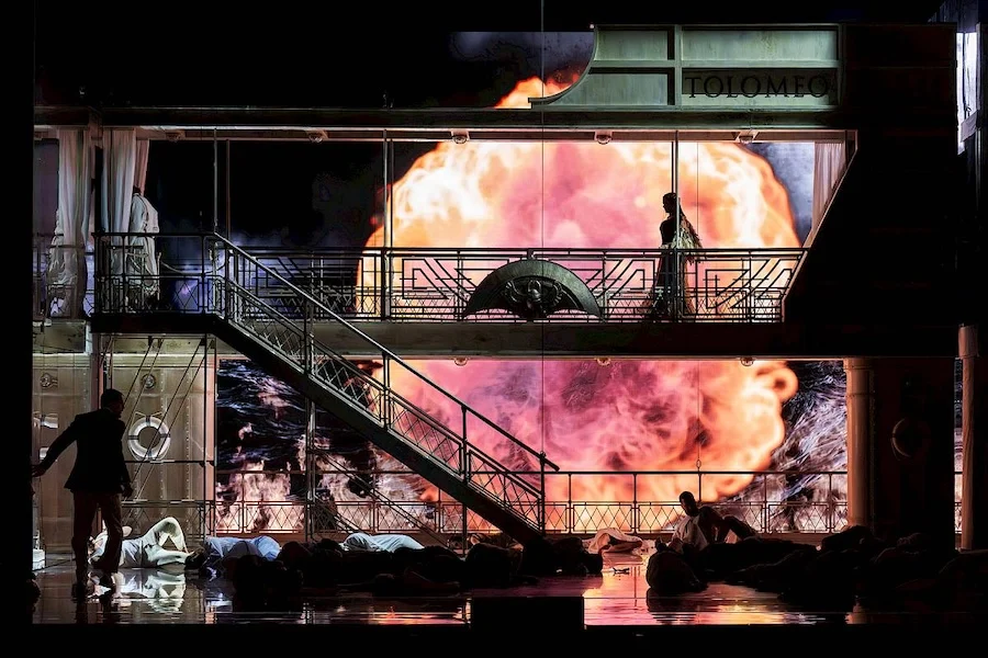 Scenografia in penombra con un'enorme esplosione arancione sullo schermo e corpi stesi a terra – Giulio Cesare in Egitto all'Opéra de Monte-Carlo, Principato di Monaco, 2025 – scenografia d'opera