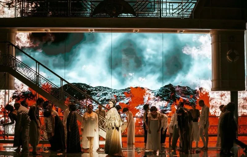 Coro in costumi chiari davanti alla proiezione di un mare di fuoco e nuvole scure – Giulio Cesare in Egitto all'Opéra de Monte-Carlo, Principato di Monaco, 2025 – scenografia d'opera