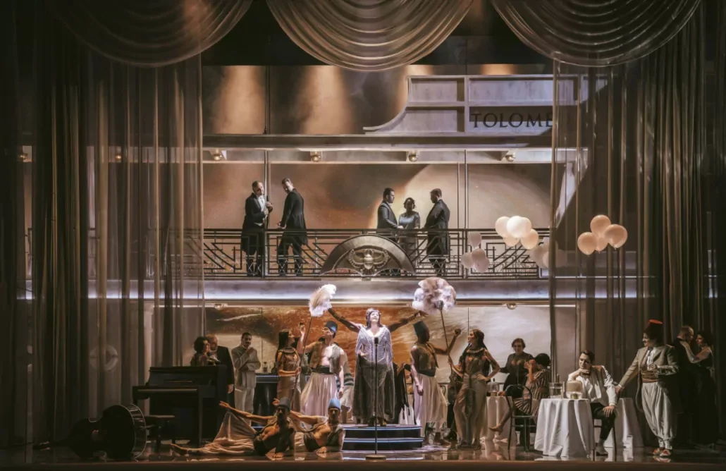 Cantante su un piccolo palco sotto tende drappeggiate, con ballerini, tavolini da caffè e uomini su un balcone sopra – Giulio Cesare in Egitto all'Opéra de Monte-Carlo, Principato di Monaco, 2025 – scenografia d'opera