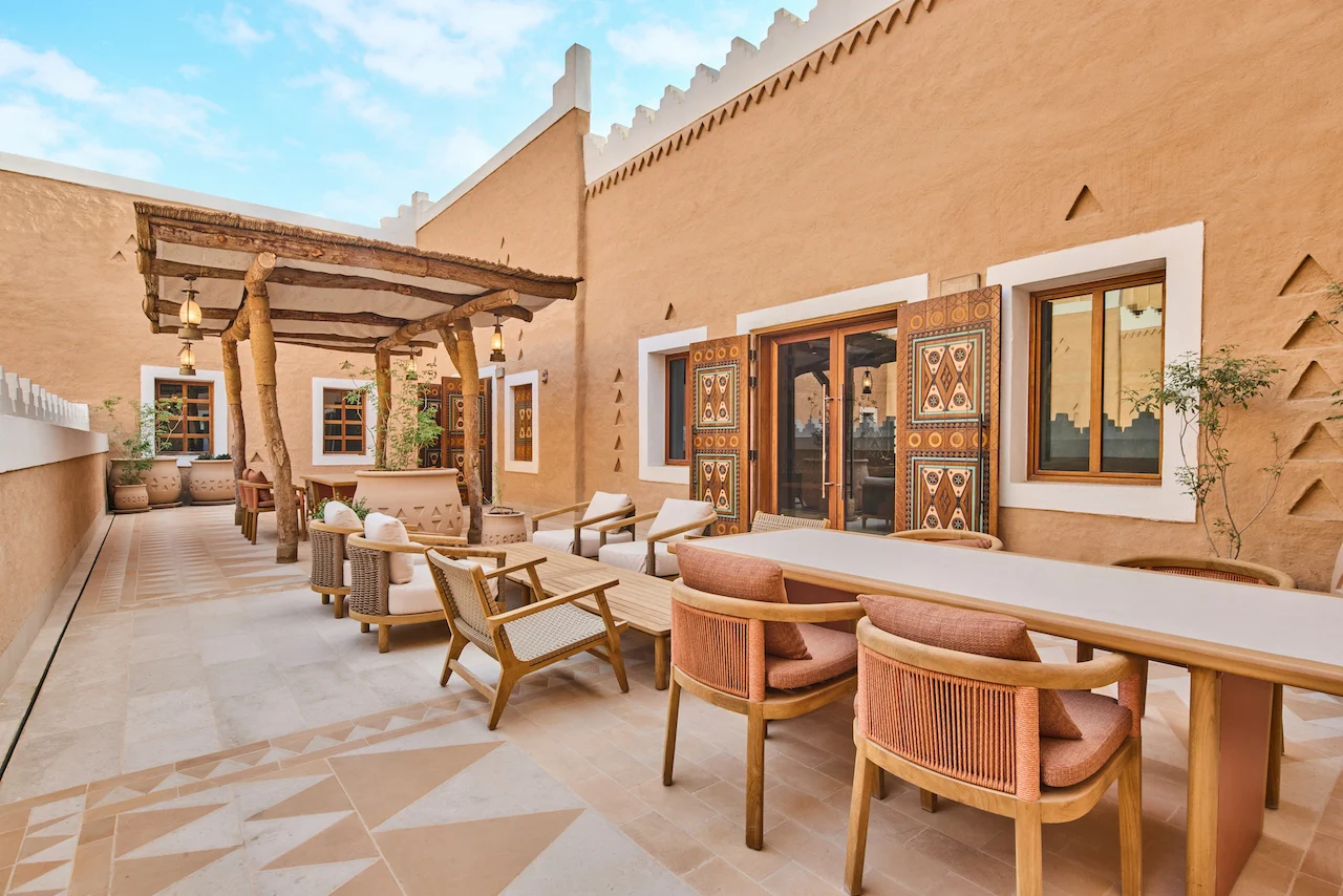 Terrazza esterna con pergola in legno, tavolo da pranzo e sedute lounge accanto a muri in terra cruda con porte dipinte – Sede del MOC, Diriyah, Arabia Saudita, 2026 – interior design