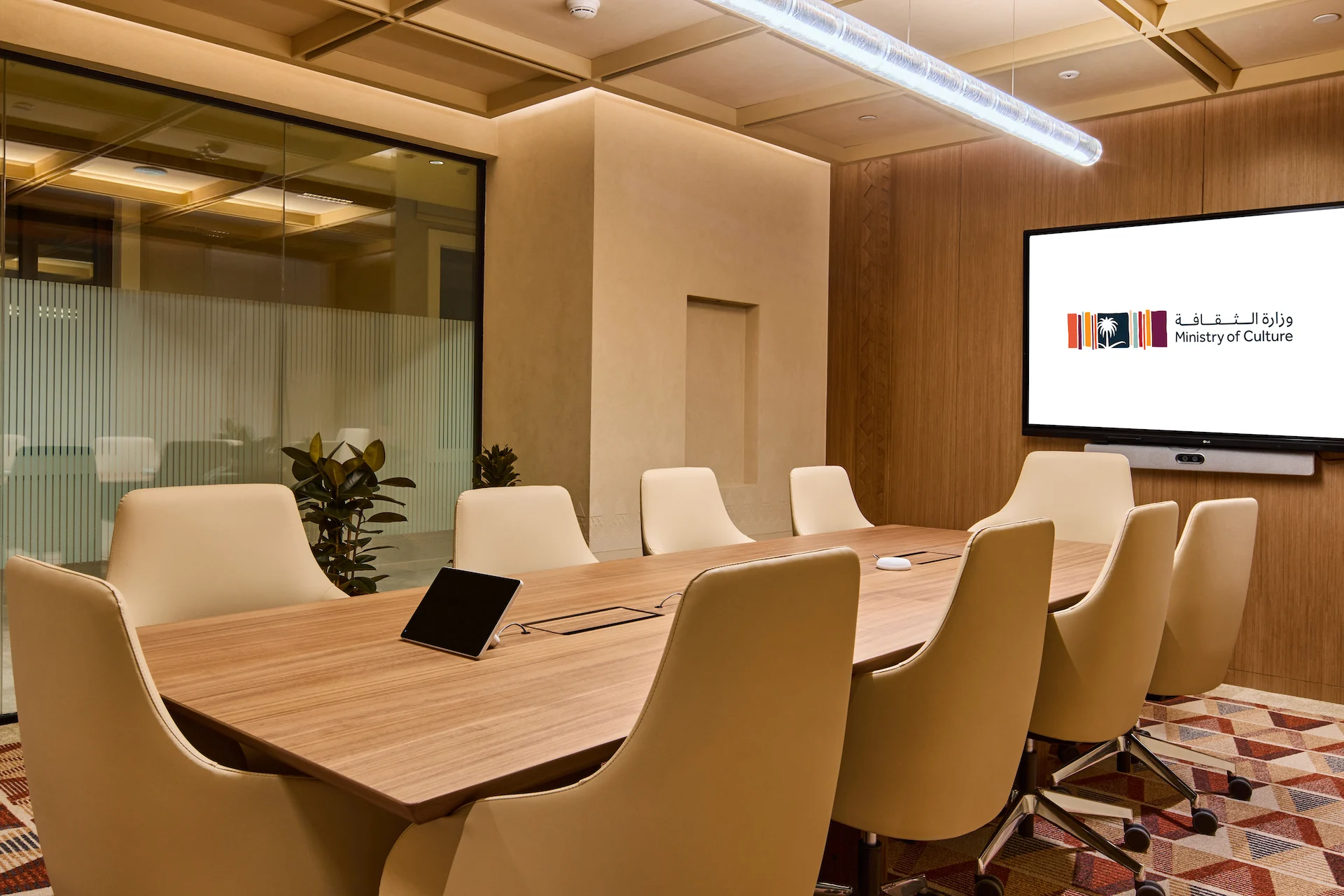 Sala riunioni con un lungo tavolo in legno, sedie color crema, parete rivestita in legno e uno schermo con il logo del Ministero – Sede del MOC, Diriyah, Arabia Saudita, 2026 – interior design