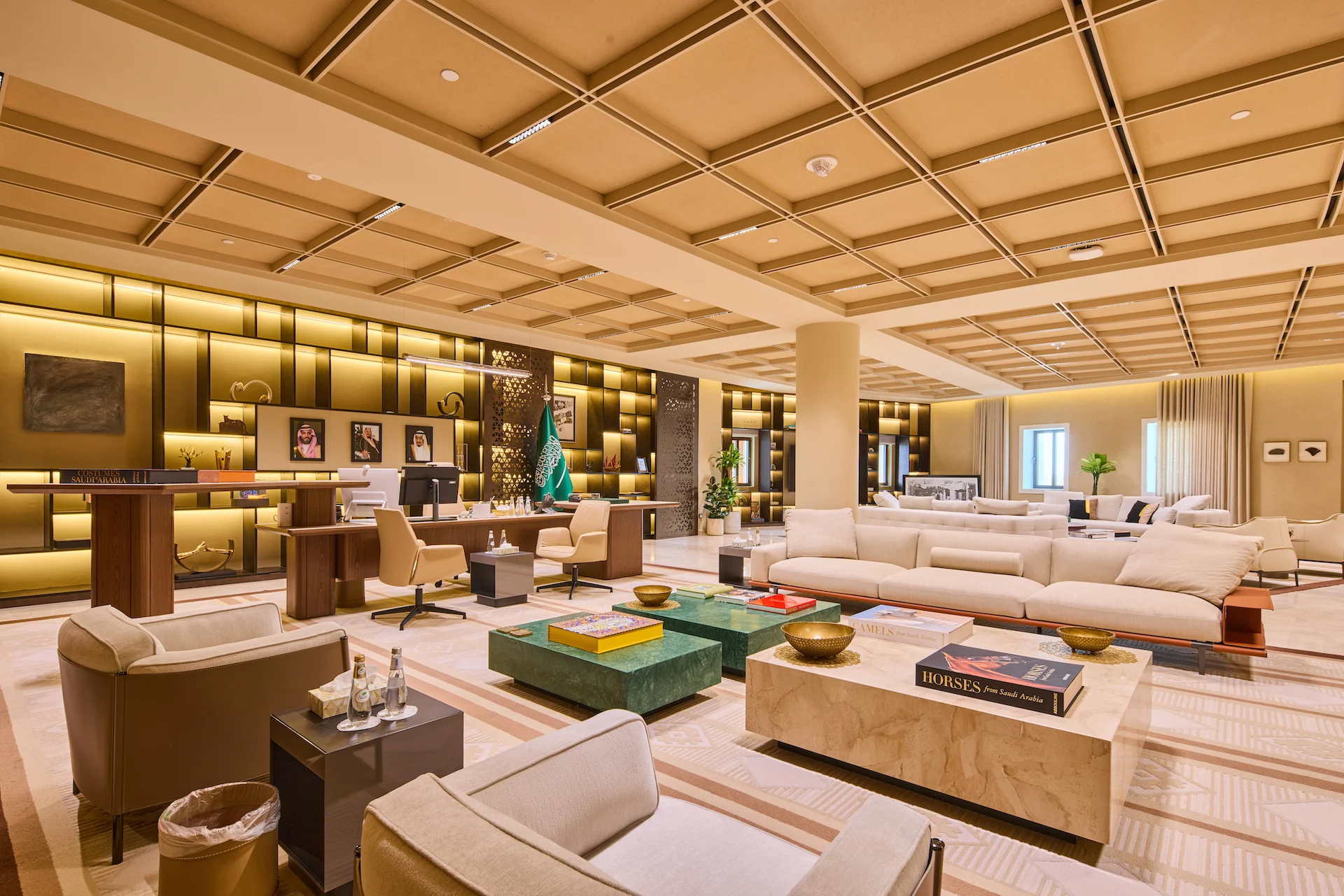Ufficio direzionale open space e lounge sotto un soffitto a cassettoni, con scaffali retroilluminati, divani e tavolini in marmo verde – Sede del MOC, Diriyah, Arabia Saudita, 2026 – interior design