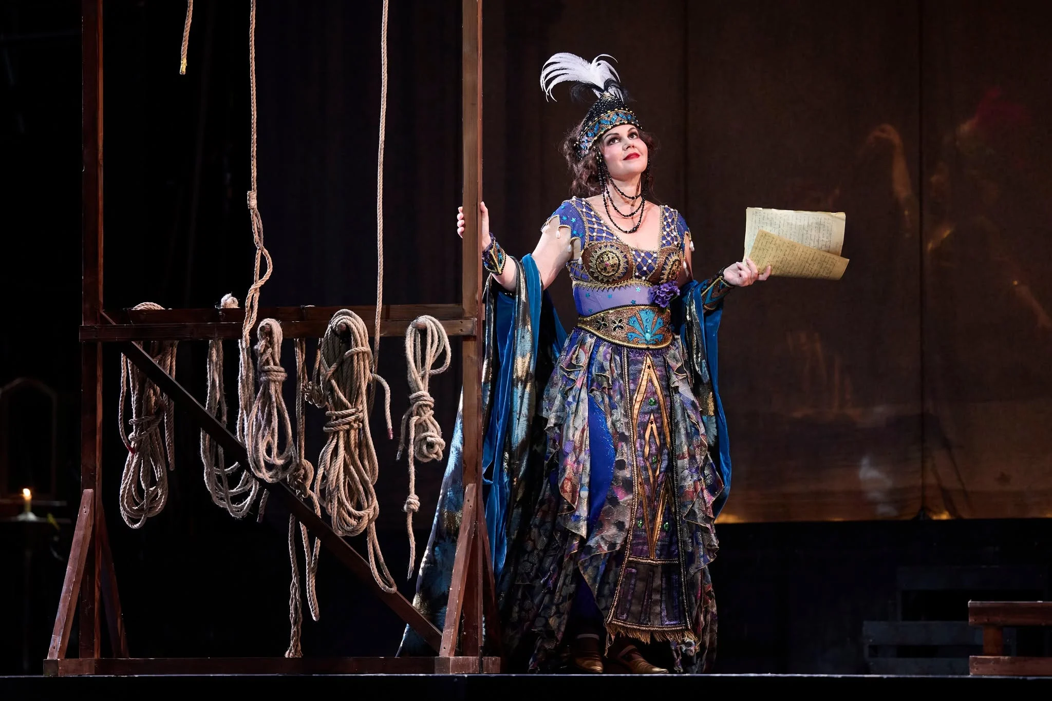 Cantante in costume blu ingioiellato e copricapo di piume con una lettera in mano, accanto a una cornice con funi appese – Adriana Lecouvreur all'Opéra de Monte-Carlo, Principato di Monaco, 2026 – scenografia d'opera