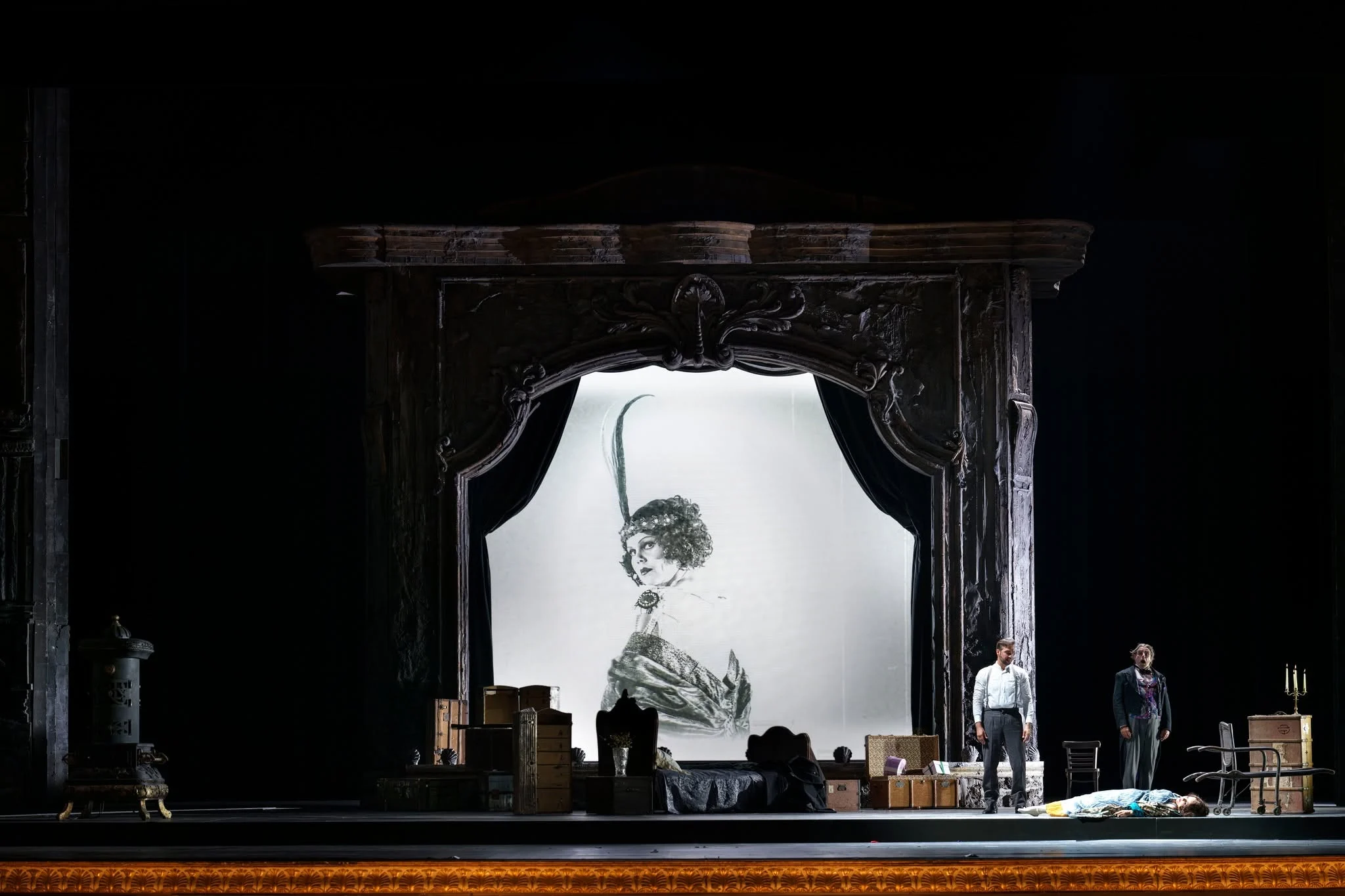 Portale scenico intagliato che inquadra un grande ritratto a schizzo di una diva, bauli e artisti davanti – Adriana Lecouvreur all'Opéra de Monte-Carlo, Principato di Monaco, 2026 – scenografia d'opera