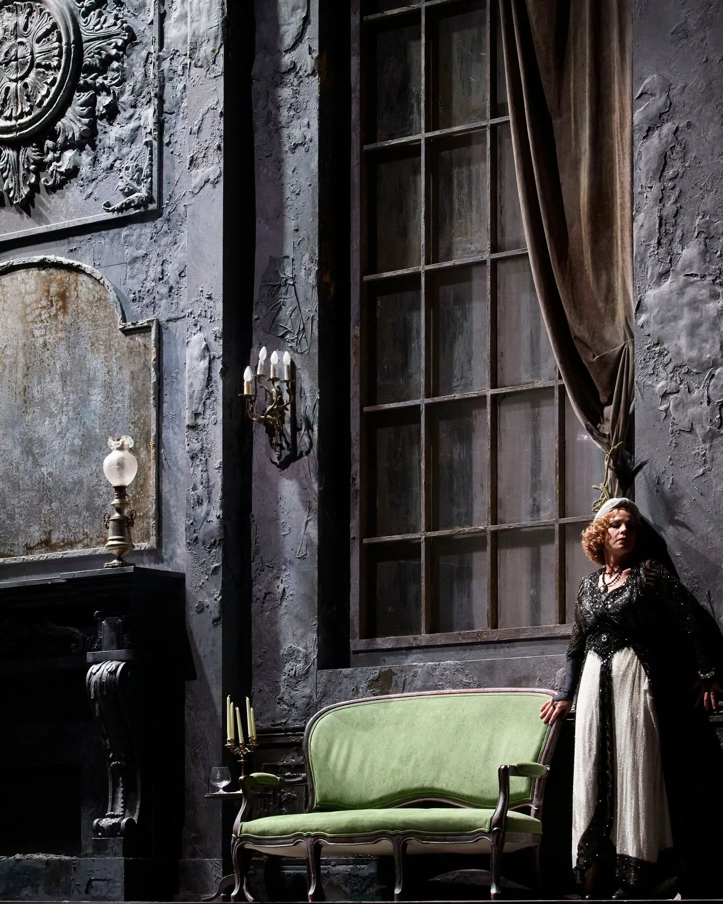 Cantante in pizzo nero accanto a un divanetto verde, in un salotto grigio consunto con un'alta finestra – Adriana Lecouvreur all'Opéra de Monte-Carlo, Principato di Monaco, 2026 – scenografia d'opera