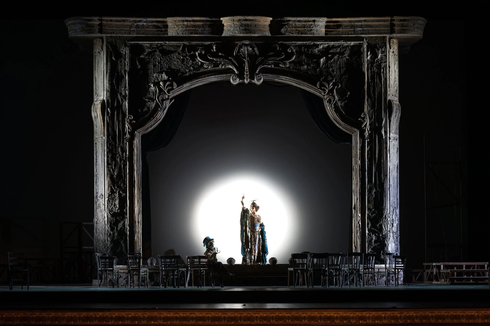 Cantante nella luce bianca di un occhio di bue al centro di un portale argentato intagliato, sedie allineate sul palco buio – Adriana Lecouvreur all'Opéra de Monte-Carlo, Principato di Monaco, 2026 – scenografia d'opera