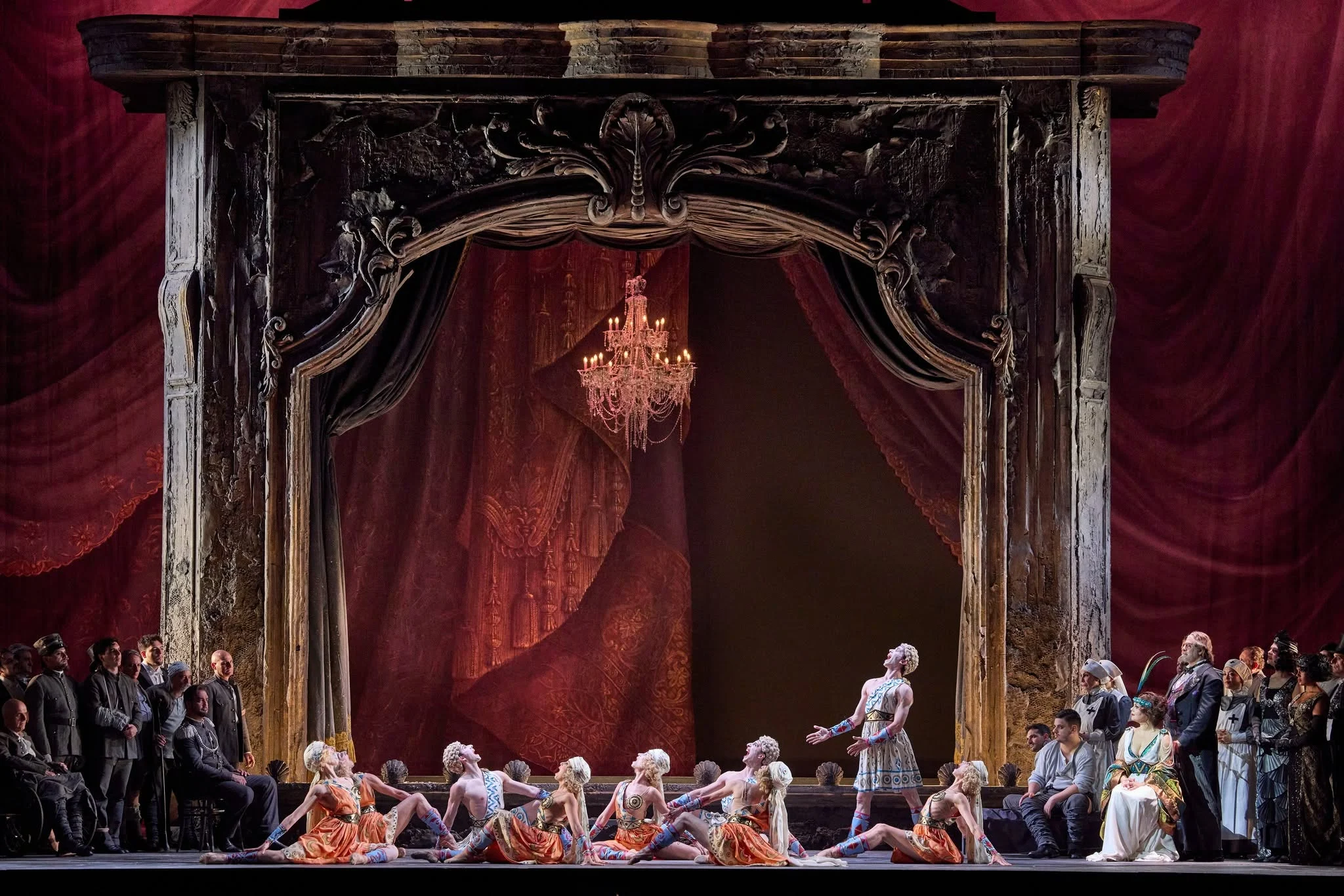 Ballerini in costumi arancioni sotto un lampadario, incorniciati dal portale intagliato e da drappi di velluto rosso – Adriana Lecouvreur all'Opéra de Monte-Carlo, Principato di Monaco, 2026 – scenografia d'opera