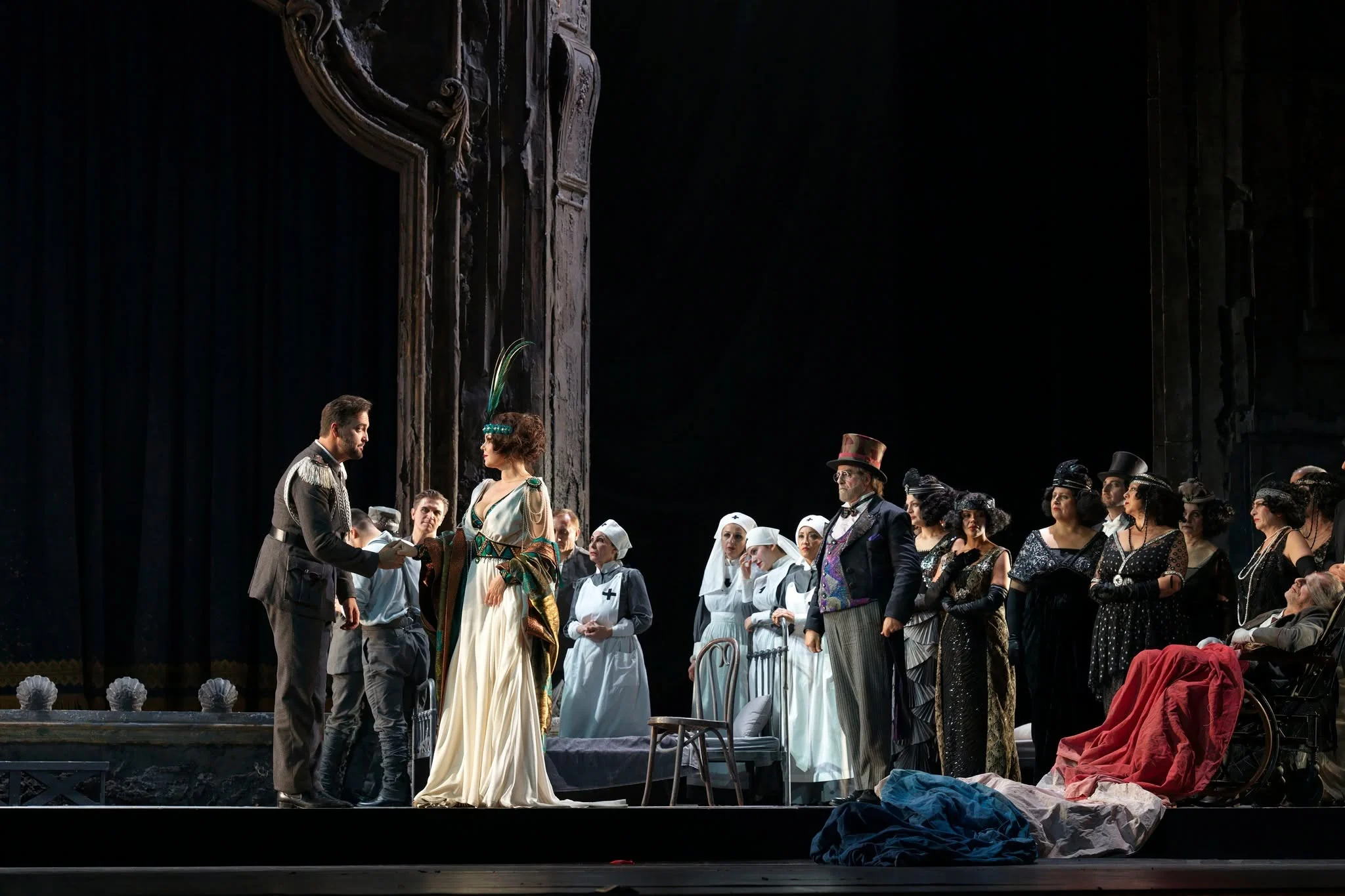 Solisti e coro in costumi d'epoca con infermiere in bianco, riuniti accanto al portale intagliato – Adriana Lecouvreur all'Opéra de Monte-Carlo, Principato di Monaco, 2026 – scenografia d'opera