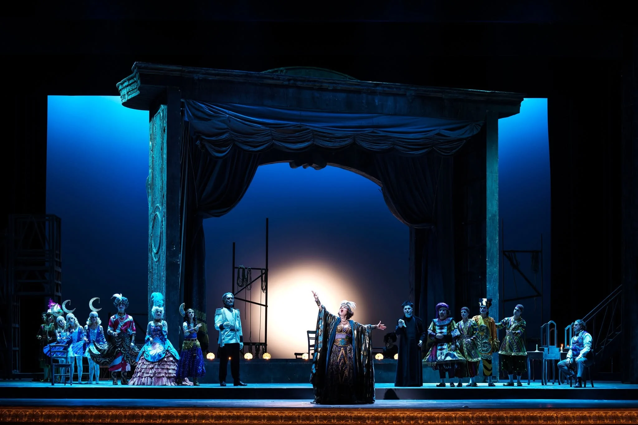 Adriana Lecouvreur al San Carlo: cantante davanti a una cornice scenica drappeggiata, interpreti in costume in luce blu intensa – Adriana Lecouvreur all'Opéra de Monte-Carlo, Principato di Monaco, 2026 – scenografia d'opera