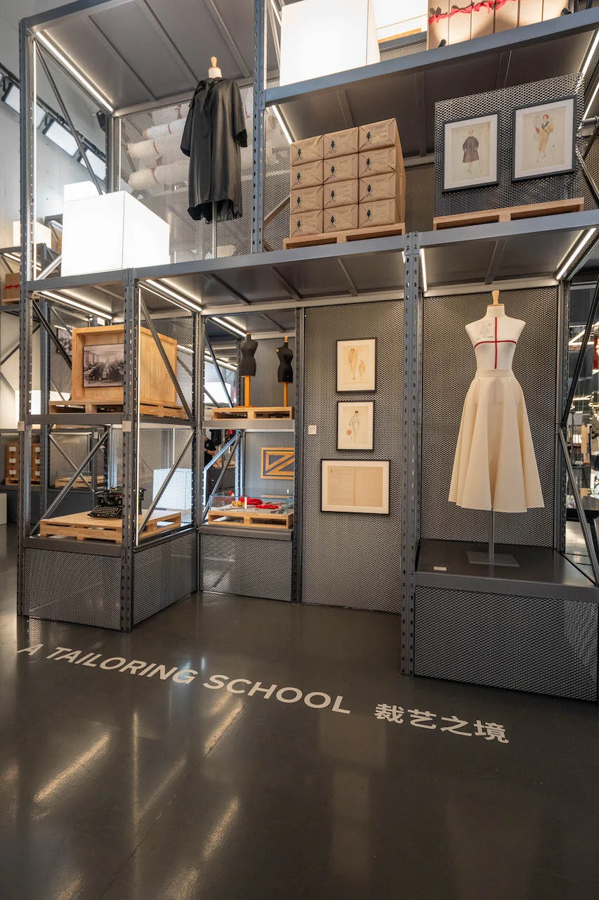 Sezione A Tailoring School: busto sartoriale bianco, schizzi incorniciati e casse di legno su scaffalature metalliche – Max Mara The Max! al Long Museum, Shanghai, Cina, 2026 – allestimento espositivo