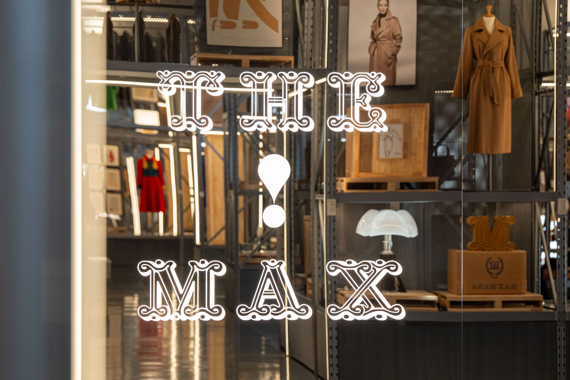 Il titolo della mostra The Max! su una porta di vetro, dietro i cappotti Max Mara esposti su scaffalature metalliche – Max Mara The Max! al Long Museum, Shanghai, Cina, 2026 – allestimento espositivo