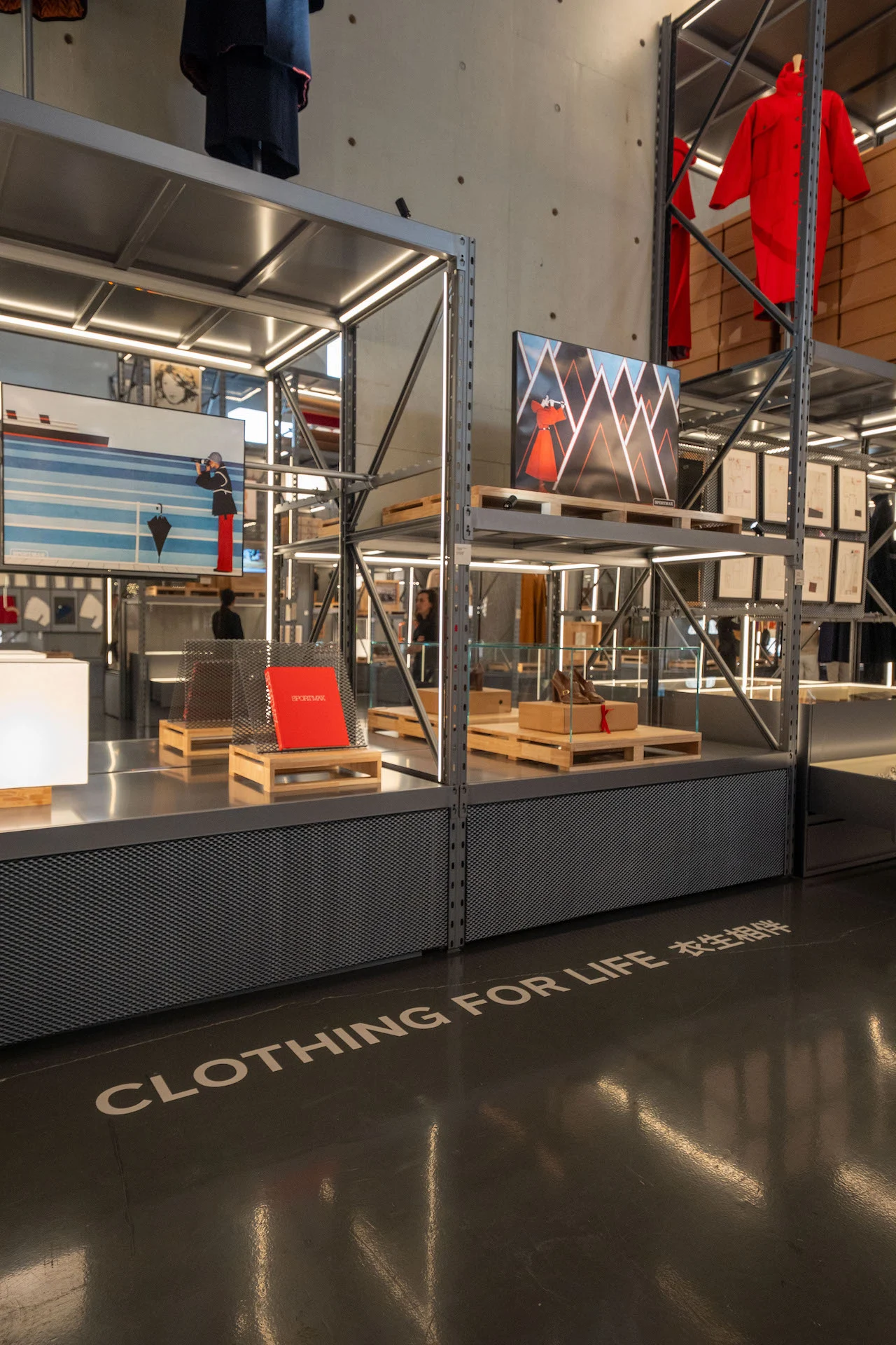 Sezione Clothing for Life: scaffali metallici con capi rossi, opere d'arte e libri su pallet di legno – Max Mara The Max! al Long Museum, Shanghai, Cina, 2026 – allestimento espositivo