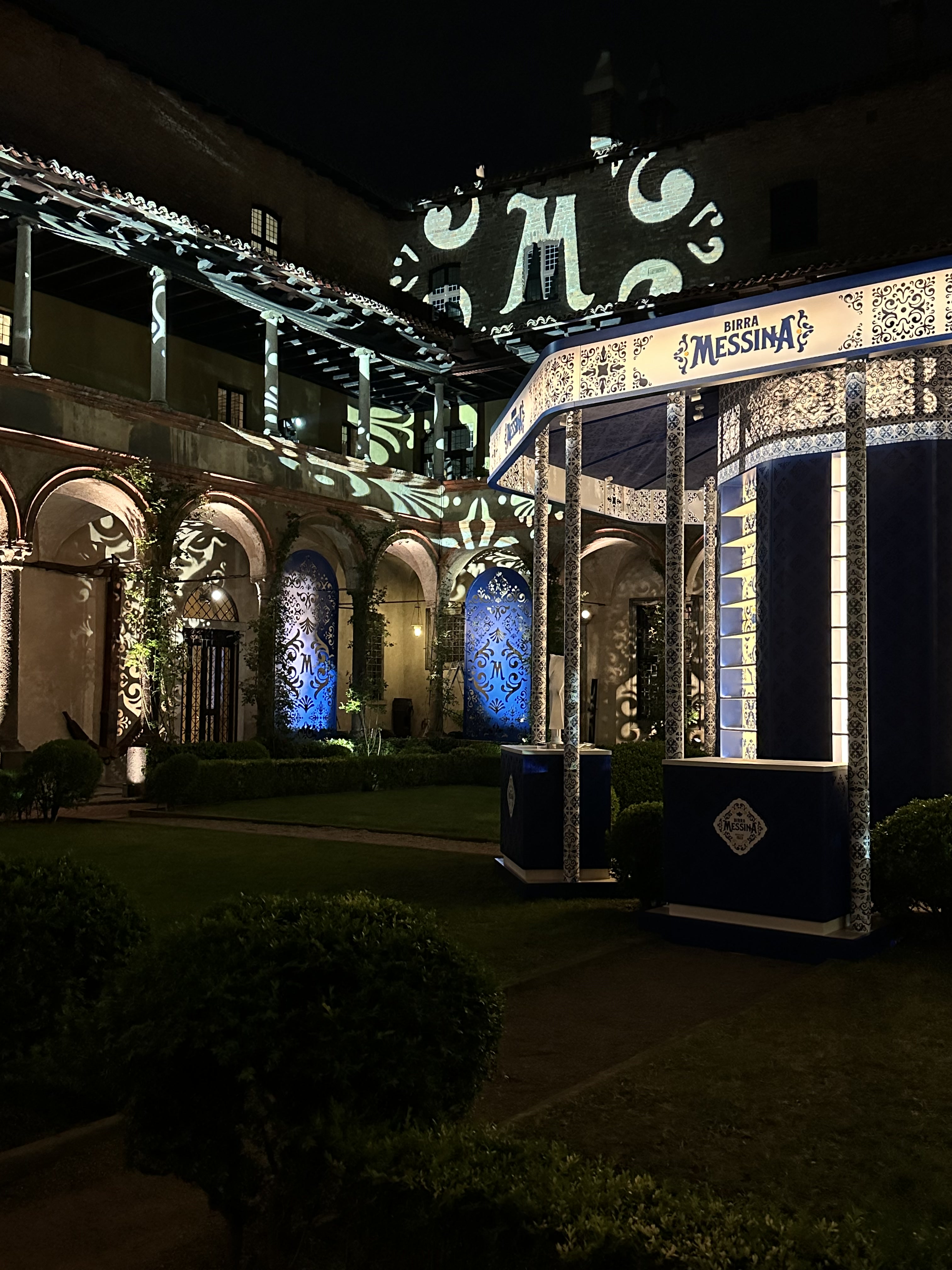 Cortile di notte con un chiosco Birra Messina e proiezioni blu a motivi sulla facciata porticata – Birra Messina Bar Meraviglia al Fuorisalone, Milano, Italia, 2024 – progettazione di eventi