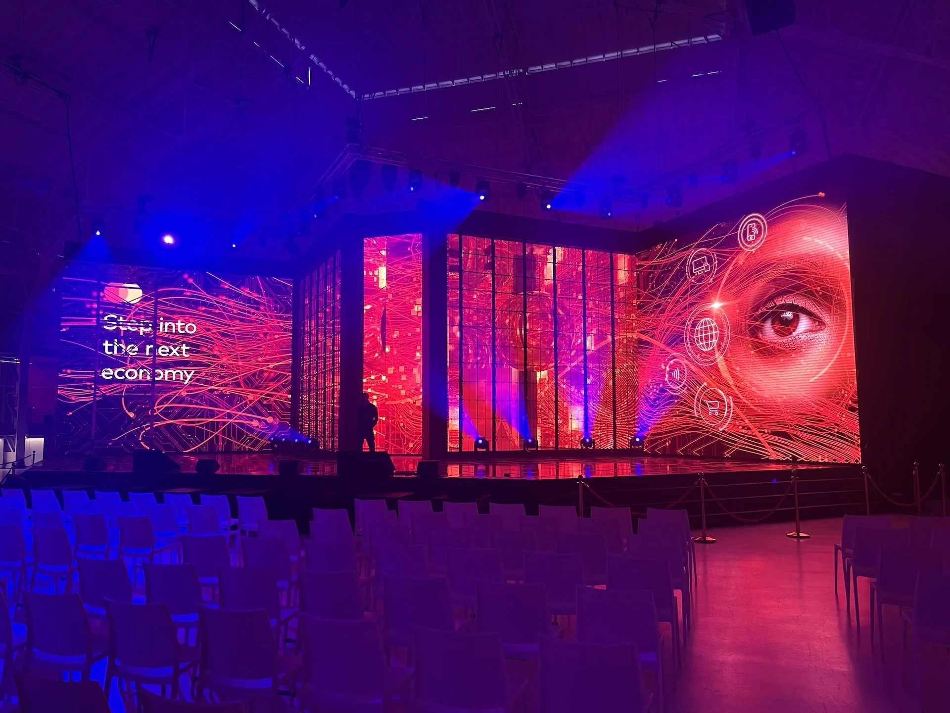 Palco con pareti LED con la scritta Step into the next economy, la grafica di un occhio gigante e file di sedie bianche – Mastercard Innovation Forum 2023, Milano, Italia – progettazione di eventi